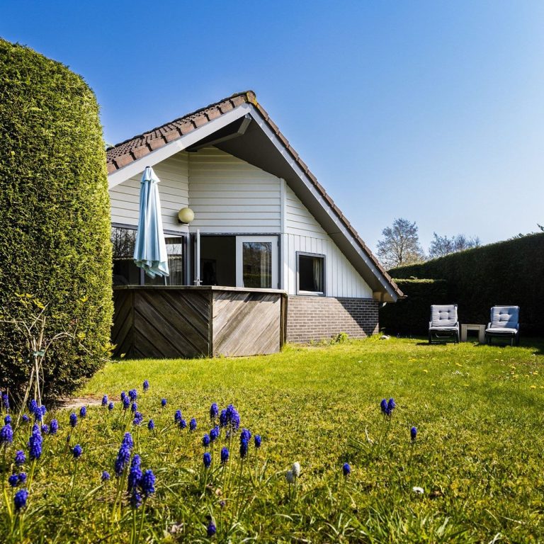 Witte vakantiewoning met tuin, blauwe lucht en paarse bloemen in het gras.