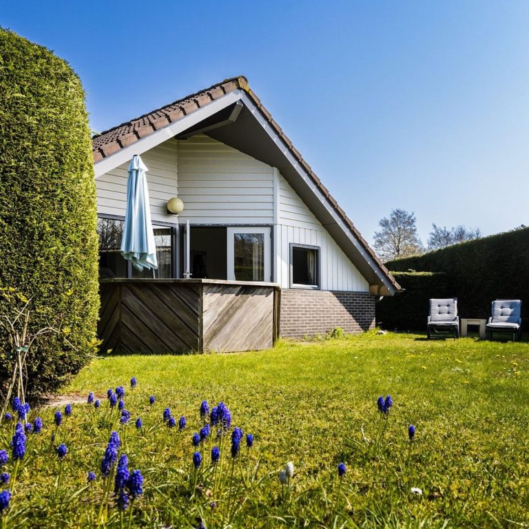 Zeebungalow op Terschelling Wit huis met tuin, omringd door groene hagen en paarse bloemen in de voorgrond.